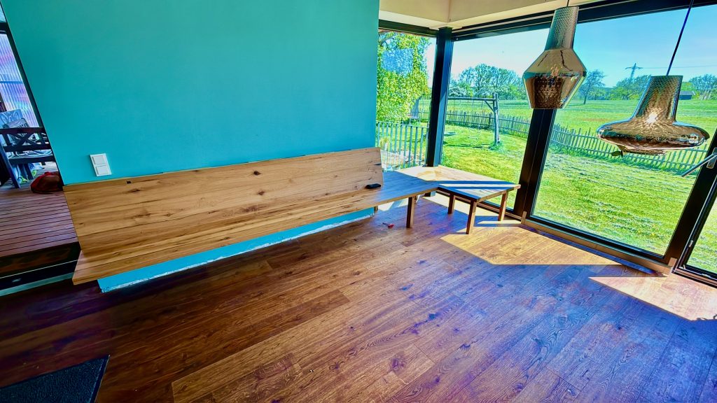 Blick in einen lichtdurchfluteten Wohnraum mit türkiser Wandfarbe und raumhohen Glasfronten. Entlang der Wand erstreckt sich eine wandmontierte Eckbank aus geölter Altholz-Eiche mit sichtbarer Maserung und natürlichen Ästen. Die Bank geht in einen freistehenden Esstisch über. Durch die Fenster ist eine grüne Wiese mit Bäumen zu sehen. Im Vordergrund ein dunkler Eichenparkett, rechts im Bild eine kupferfarbene Hängeleuchte.